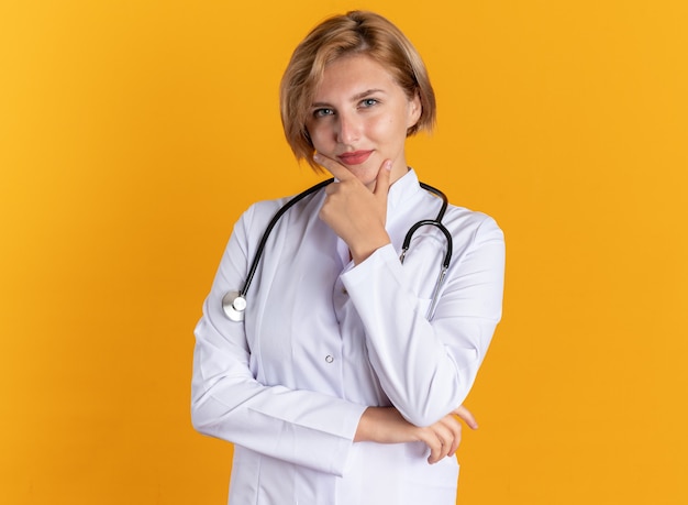 Tevreden jonge vrouwelijke arts die medisch gewaad met stethoscoop draagt, greep kin geïsoleerd op oranje muur on