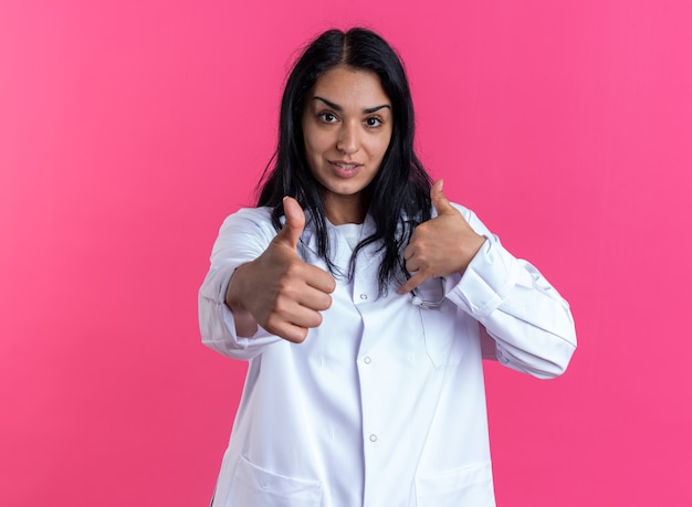 Tevreden jonge vrouwelijke arts die medische mantel draagt met een stethoscoop die duimen laat zien geïsoleerd op roze muur