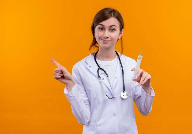 Tevreden jonge vrouwelijke arts die medische mantel en stethoscoop draagt ?? die spuit en naar de linkerkant op geïsoleerde oranje ruimte met exemplaarruimte richt