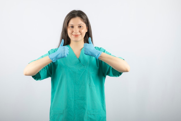 Tevreden jonge vrouwelijke arts in medische handschoenen die duimen opdagen.