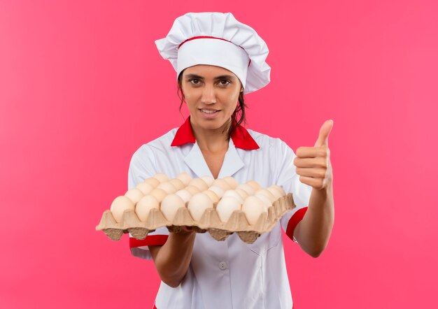 tevreden jonge vrouwelijke kok die chef-kok uniform draagt die partij eieren herthumb omhoog op geïsoleerde roze muur met exemplaarruimte houdt