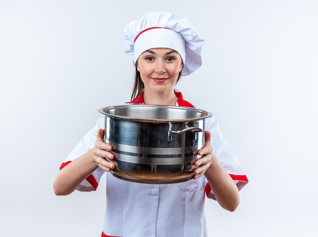 tevreden jonge vrouwelijke kok die een chef-kokuniform draagt die een steelpan houdt die op witte muur wordt geïsoleerd