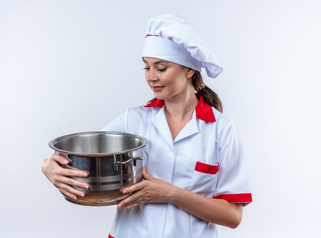 Tevreden jonge vrouwelijke kok die een chef-kokuniform draagt en naar een steelpan kijkt die op een witte muur wordt geïsoleerd