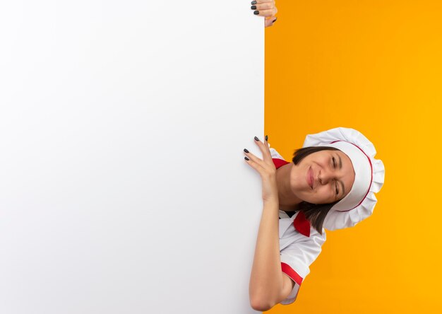 Tevreden jonge vrouwelijke kok in eenvormige chef-kok die zich achter witte muur houdt en hand erop legt met gesloten ogen die op oranje muur worden geïsoleerd