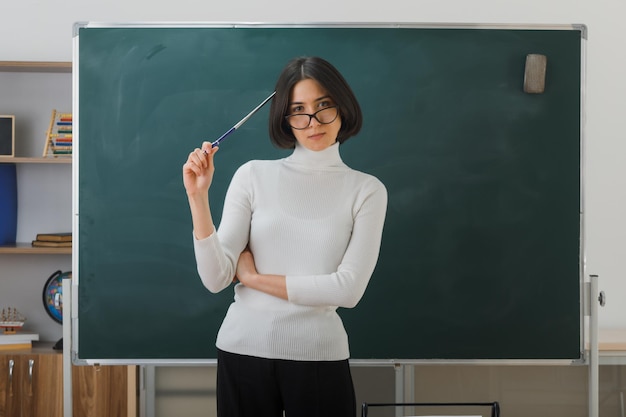 Tevreden jonge vrouwelijke leraar die een bril draagt die vooraan op het schoolbord staat met de aanwijzer op het hoofd in de klas