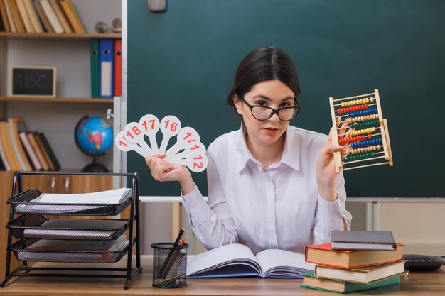 tevreden jonge vrouwelijke leraar met een bril die een telraam vasthoudt met een nummerventilator die aan het bureau zit met schoolhulpmiddelen in de klas