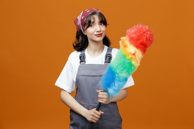 Tevreden jonge vrouwelijke schoonmaker die uniform en bandana draagt die plumeau met beide handen houdt die camera bekijken die op oranje achtergrond wordt geïsoleerd