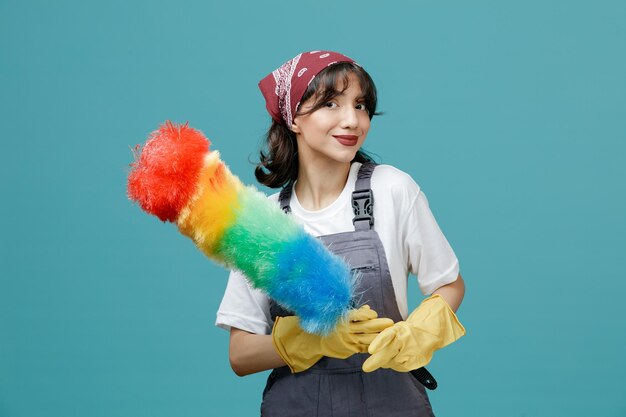 Tevreden jonge vrouwelijke schoonmaker die uniforme bandana en rubberen handschoenen draagt met plumeau met beide handen kijkend naar camera geïsoleerd op blauwe achtergrond
