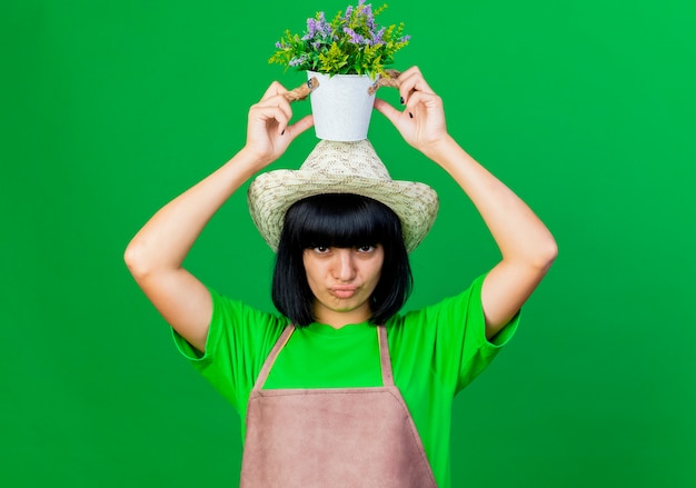 Tevreden jonge vrouwelijke tuinman in uniform met tuinhoed houdt bloempot boven het hoofd