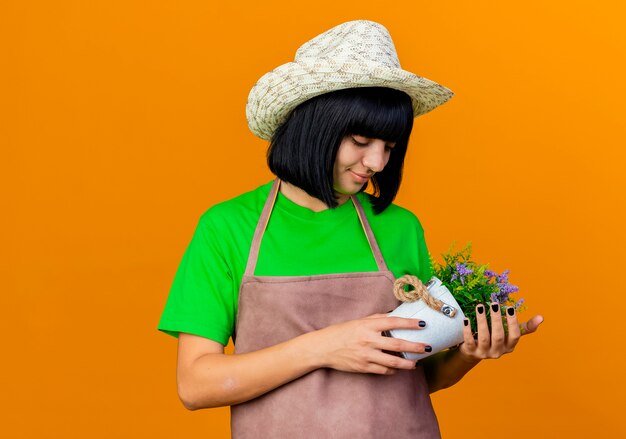 Tevreden jonge vrouwelijke tuinman in uniform met tuinhoed houdt bloempot vast