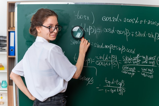 Tevreden jonge vrouwelijke wiskundeleraar met een bril die achter het zicht voor het schoolbord staat