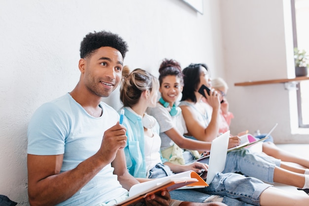 Tevreden jongeman met boeken en leerboeken met glimlach opzoeken, terwijl zijn klasgenoten iets bespreken. Binnenportret van studenten die zich voorbereiden op het examen.