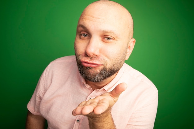 Gratis foto tevreden kale man van middelbare leeftijd die roze t-shirt draagt die kusgebaar toont dat op groen wordt geïsoleerd