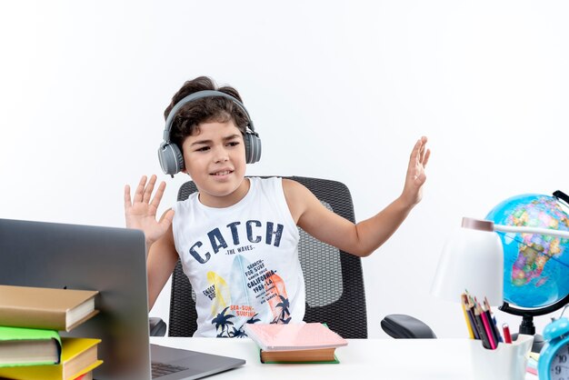 Tevreden kleine schooljongen die hoofdtelefoons draagt die aan bureau met schoolhulpmiddelen zitten luisteren muziek die op witte achtergrond wordt geïsoleerd