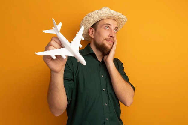 Gratis foto tevreden knappe blonde man met strandhoed legt hand op gezicht en houdt modelvliegtuig vast dat op oranje muur wordt geïsoleerd