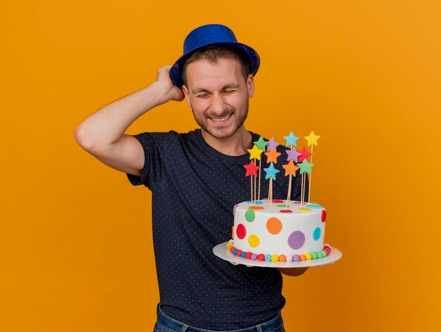 Tevreden knappe man die blauwe feestmuts draagt en houdt houdt verjaardagstaart geïsoleerd op een oranje muur met kopie ruimte