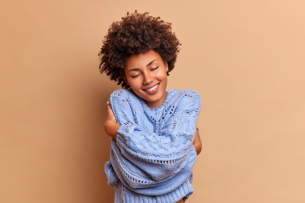 Tevreden lachende vrouw met borstelig krullend haar omhelst zichzelf met liefde geniet van zachtheid van nieuwe trui voelt comfortabel en verheugd sluit ogen tevreden geïsoleerd over beige muur