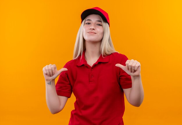 Tevreden levering jong meisje met rode t-shirt en pet wijst naar zichzelf op geïsoleerde oranje achtergrond