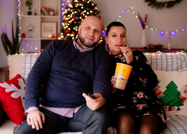 Gratis foto tevreden man en vrouw thuis met kerstmis zittend op de bank in de woonkamer man met afstandsbediening vrouw met popcorn emmer die popcorn eet allebei tv kijken