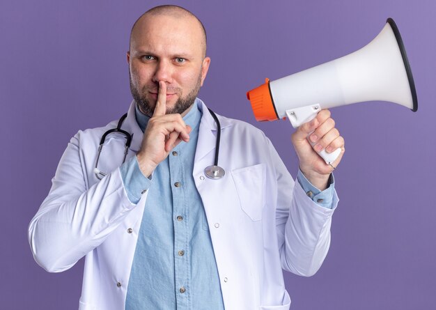 Tevreden mannelijke arts van middelbare leeftijd met een medisch gewaad en een stethoscoop die een spreker vasthoudt die een stiltegebaar doet geïsoleerd op een paarse muur