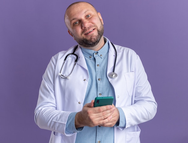 Tevreden mannelijke arts van middelbare leeftijd met medische mantel en stethoscoop met mobiele telefoon geïsoleerd op paarse muur