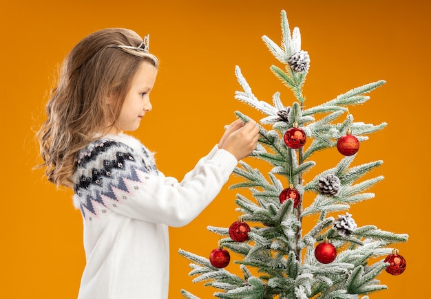 Tevreden meisje dat zich dichtbij kerstboom bevindt die tiara met slinger op de boom van de halsholding op oranje achtergrond draagt
