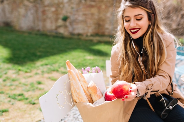 Gratis foto tevreden met winkelend meisje dat met grote glimlach haar aankopen bekijkt. aantrekkelijke jonge vrouw lachen en eten vouwen in de papieren zak zittend in het park.