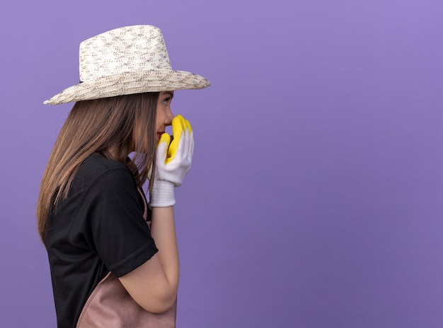 Gratis foto tevreden, mooie blanke vrouwelijke tuinman die een tuinhoed en handschoenen draagt, staat zijwaarts en houdt handen dicht bij de mond