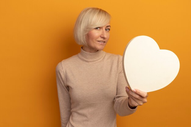 Tevreden mooie blonde Slavische vrouw houdt hartvorm kijkend naar voorzijde geïsoleerd op oranje muur