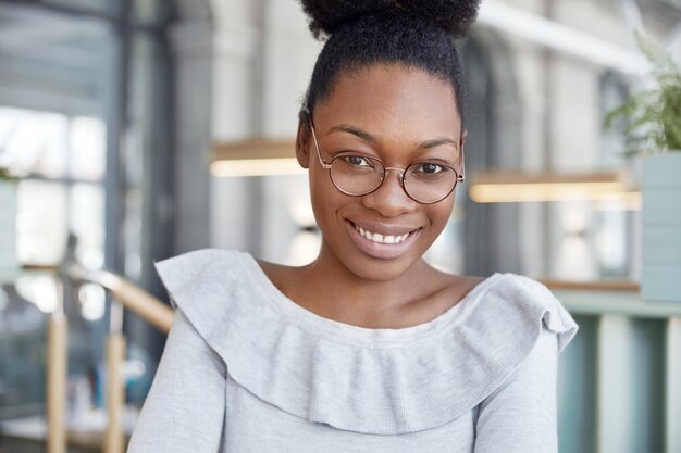 Tevreden mooie donkere vrouwelijke student met positieve uitdrukking, draagt een grote ronde bril, blij om goed cijfer te ontvangen op de universiteit, vormt binnen.