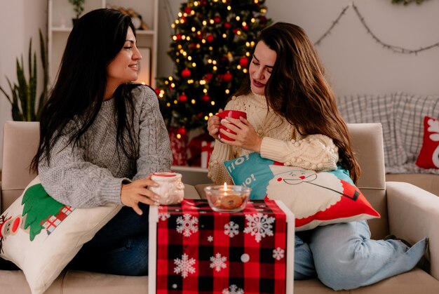 tevreden mooie jonge meisjes houden en kijken naar bekers zittend op fauteuils en genieten van kersttijd thuis