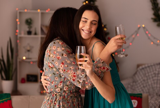 tevreden mooie jonge meisjes omhelzen elkaar en houden glazen champagne thuis genieten van kersttijd