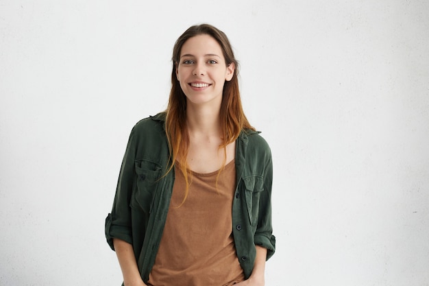 Gratis foto tevreden mooie vrouw met geverfd haar, donkere ogen en gezonde huid, gekleed in een bruin t-shirt, een groen jasje, hand in hand in de zakken glimlachend terwijl ze poseert tegen een witte betonnen muur. mensen en levensstijl