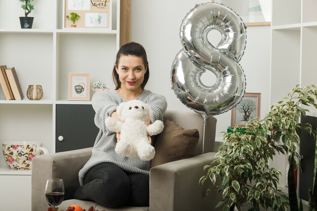 Tevreden mooie vrouw op gelukkige vrouwendag die teddybeer uithoudt bij camera zittend op fauteuil in woonkamer