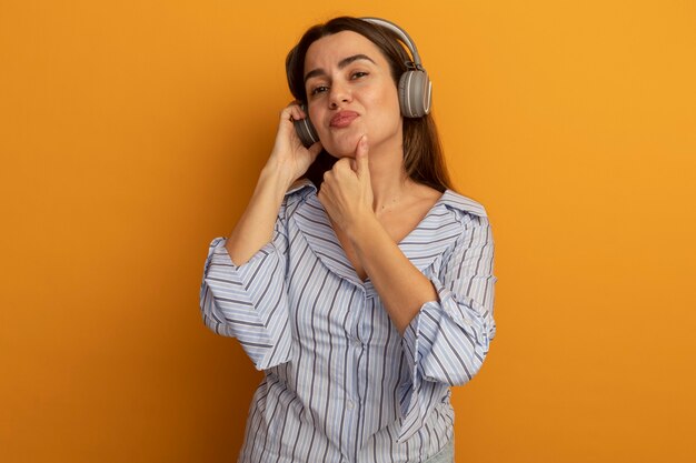 Tevreden mooie vrouw op koptelefoon houdt kin geïsoleerd op oranje muur