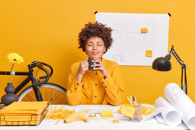 Gratis foto tevreden ontspannen vrouw architect kantoormedewerker heeft koffiepauze sluit ogen van plezier als geuren aromantische drank trekt tocht op desktop creëert schetsen