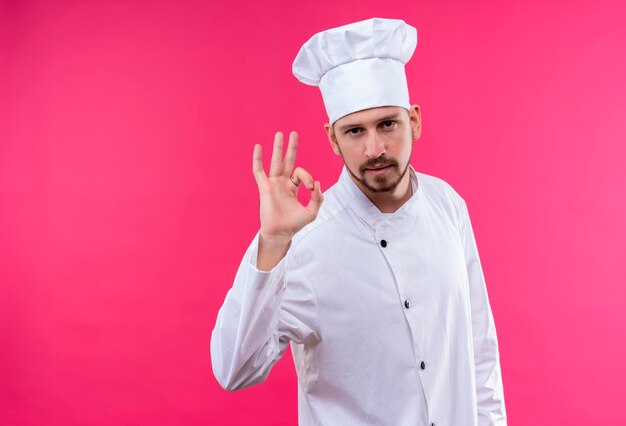 Tevreden professionele mannelijke chef-kok in wit uniform en kokhoed met ok gebaar op zoek naar vertrouwen staande over roze achtergrond