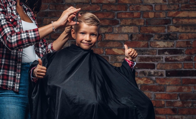 Tevreden schattige kleuterjongen toont duimen terwijl hij wordt geknipt. Kinderkapper met schaar en kam snijdt jongetje in de kamer met loft-interieur.