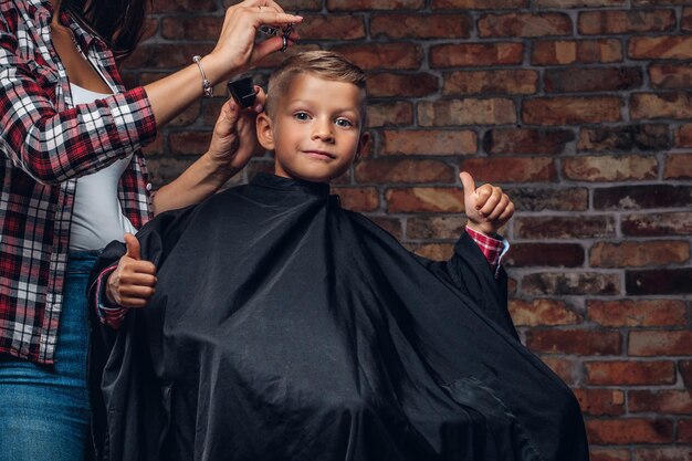 Tevreden schattige kleuterjongen toont duimen terwijl hij wordt geknipt. Kinderkapper met schaar en kam snijdt jongetje in de kamer met loft-interieur.