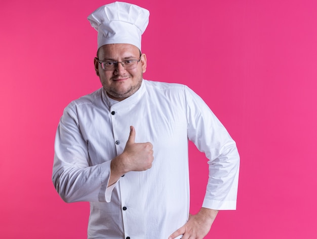 Tevreden volwassen mannelijke kok met een uniform van de chef-kok en een bril die de hand op de taille houdt en naar de voorkant kijkt met duim omhoog geïsoleerd op roze muur
