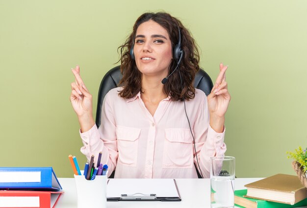 Tevreden, vrij blanke vrouwelijke callcenter-operator op een koptelefoon die aan het bureau zit met kantoorhulpmiddelen die haar vingers kruisen