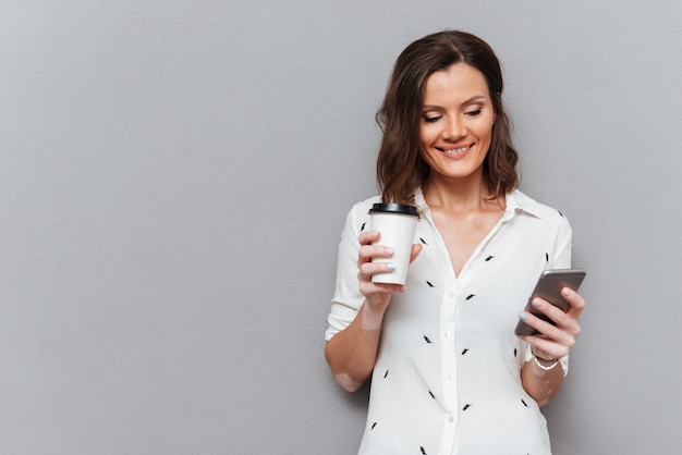 Tevreden vrouw gebruikend smartphone en houdend kop van koffie op grijs