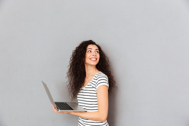 Tevreden vrouw met het krullende haar stellen met zilveren laptop die over grijze muur worden geïsoleerd die rond bekijkend iets boeiend draaien