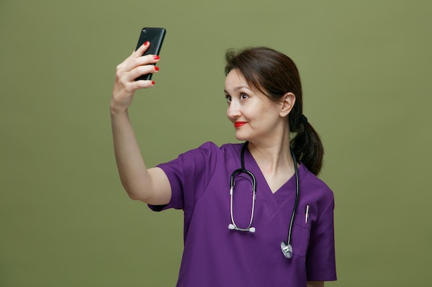 Tevreden vrouwelijke arts van middelbare leeftijd, gekleed in uniform en stethoscoop om de nek, die mobiele telefoon opheft en selfie neemt geïsoleerd op olijfgroene achtergrond