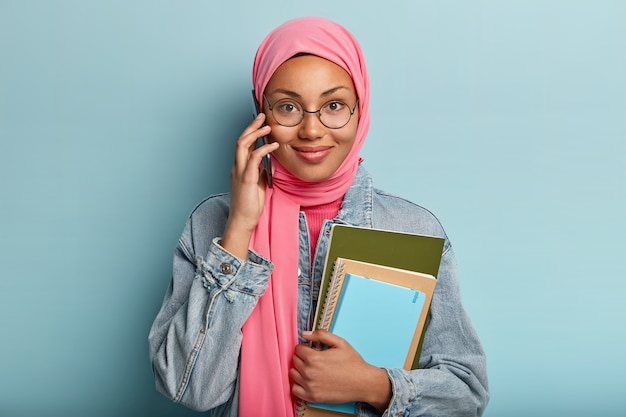 Tevreden vrouwelijke gesprekken op mobiele telefoon, besteedt vrije tijd aan roddelen, voelt zich tevreden, houdt blocnotes vast, draagt modieuze denim kleding, heeft hoofd bedekt met roze sluier, geïsoleerd over blauwe muur