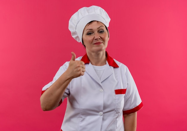 Tevreden vrouwelijke kok van middelbare leeftijd in eenvormige chef-kok haar duim omhoog op geïsoleerde roze muur met exemplaarruimte