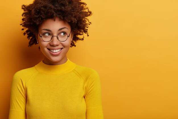 Tevreden vrouwelijke vrouw kijkt met een glimlach opzij, heeft een optimistische houding, wacht op het goede dat gebeurt, concentreert zich op mysterieuze wijze op promo-aankondiging, draagt een ronde bril, een gele trui.