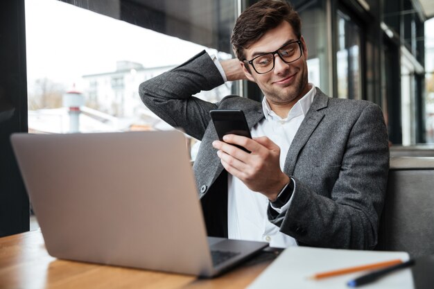 Tevreden zakenman die in oogglazen door de lijst in koffie met laptop computer zitten terwijl het gebruiken van smartphone