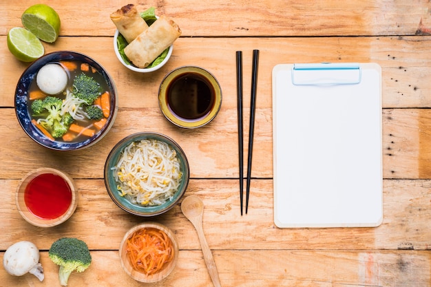 Thaise soep; loempia&#39;s; sausen en spruiten bonen met chopstick en wit papier op klembord over de houten tafel