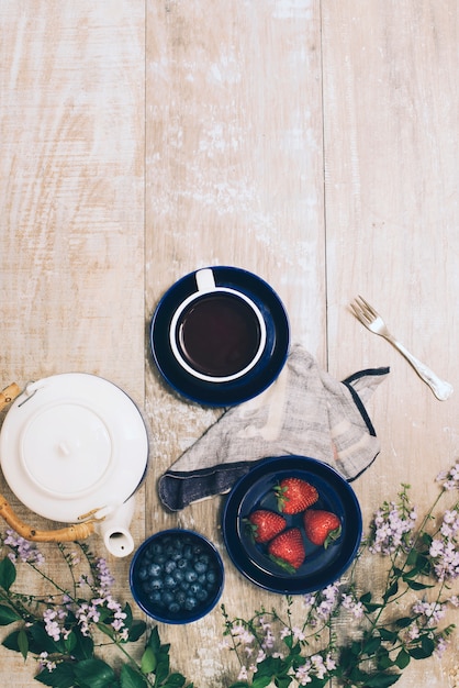 Gratis foto theepot; koffiekop; bessen; vork met servet en bloemen op houten gestructureerde achtergrond